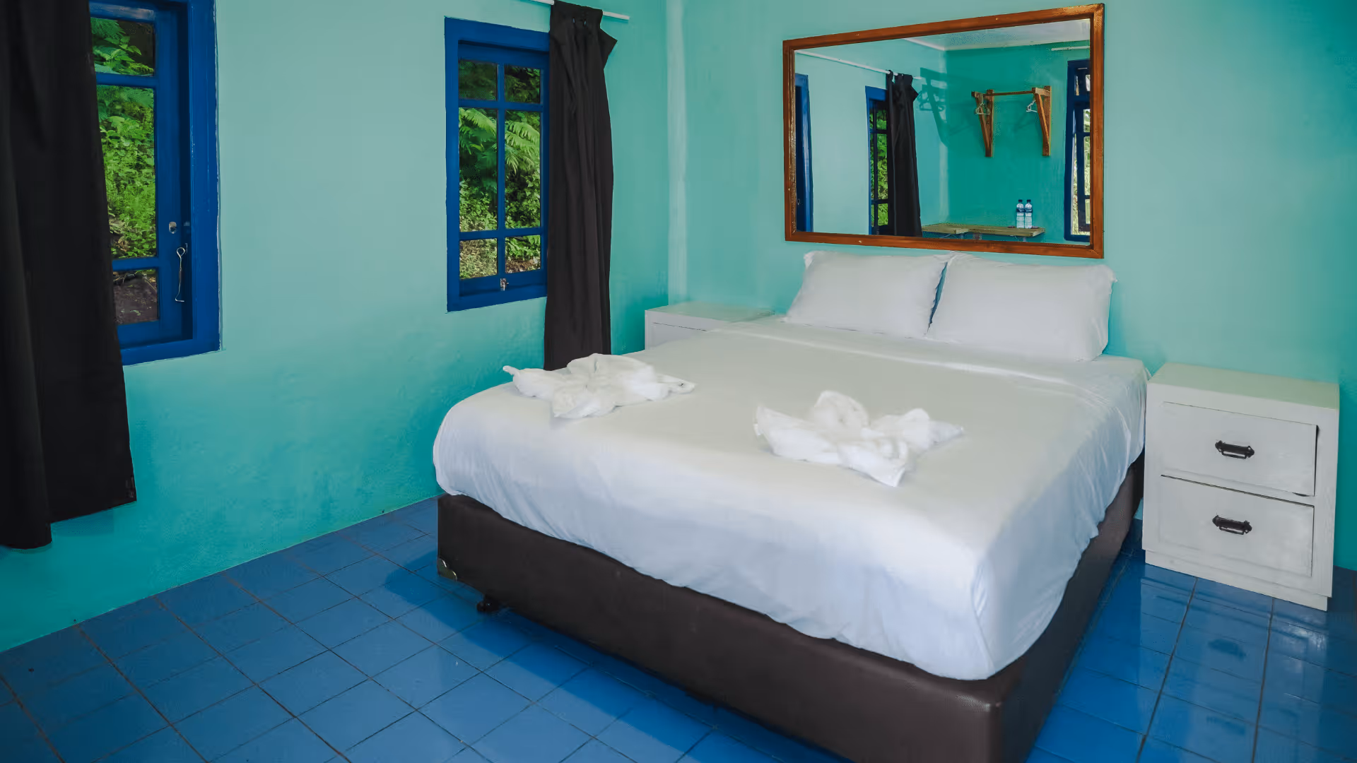 Bedroom with a large bed made with white linens, two towel swans on the bed, blue-tiled floor, turquoise walls, two blue-framed windows with dark curtains, a white nightstand, and a large wall mirror.