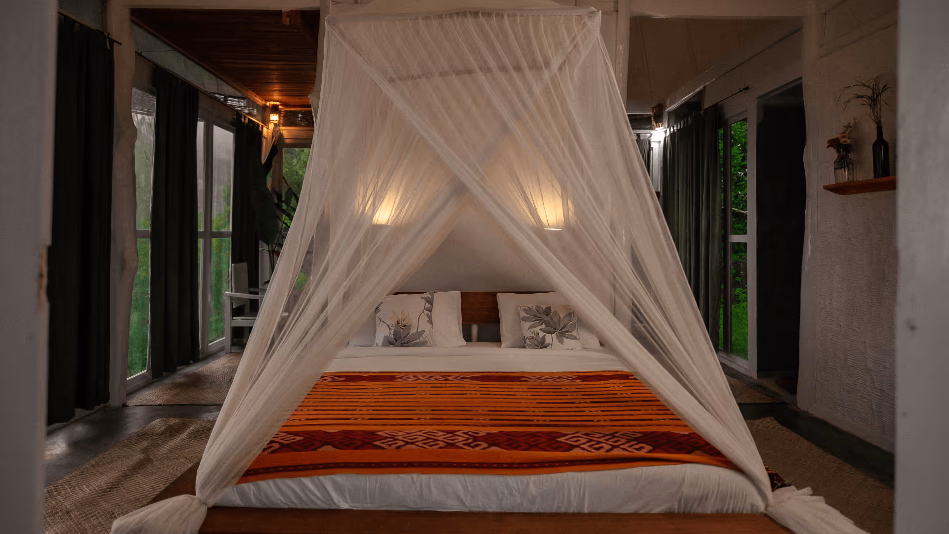 Cozy bedroom with a bed draped in a white mosquito net, orange patterned blanket, and floral pillows, surrounded by large windows with dark curtains.
