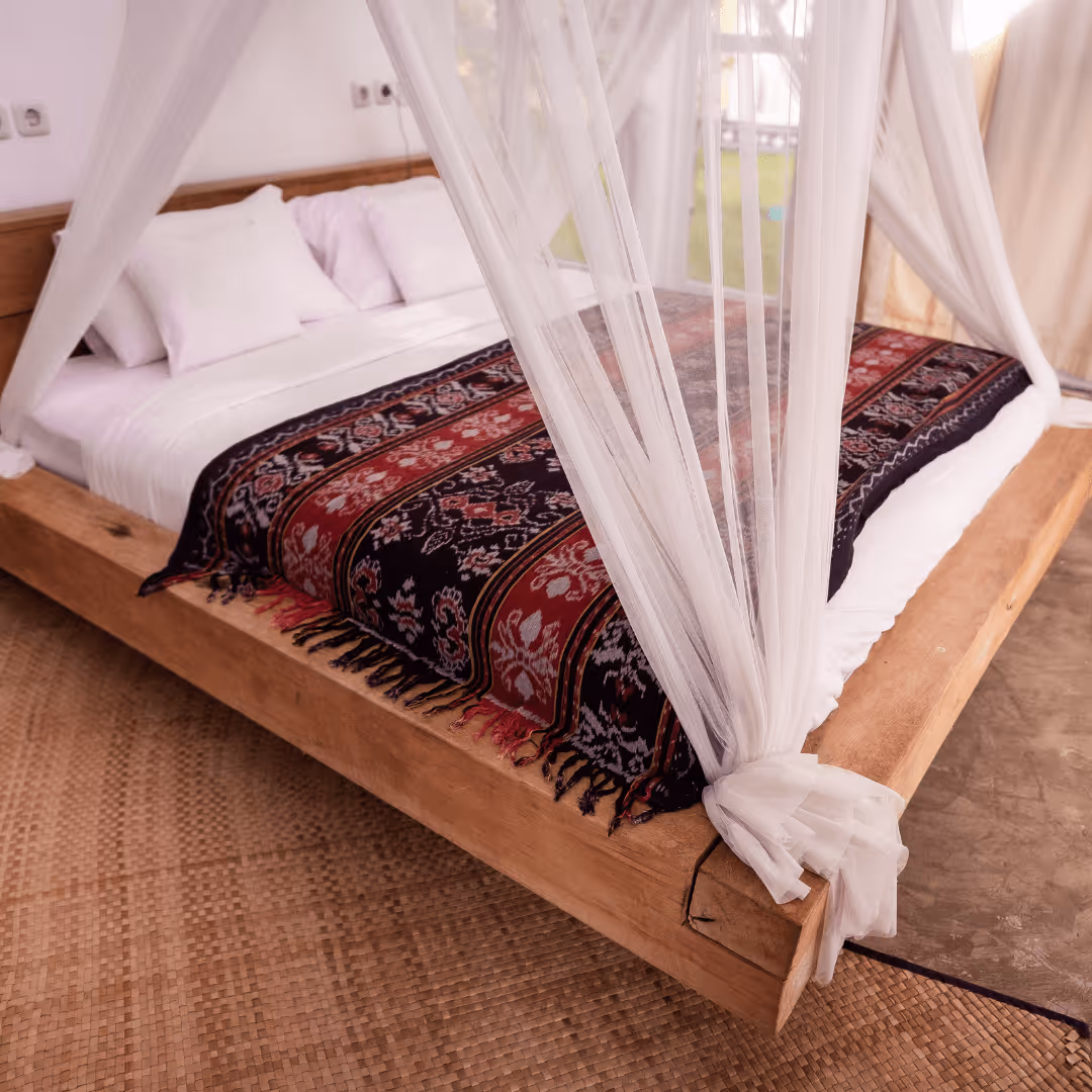 Wooden bed with white linens, pillows, a patterned red and black blanket, and sheer white canopy curtains tied at the corners.