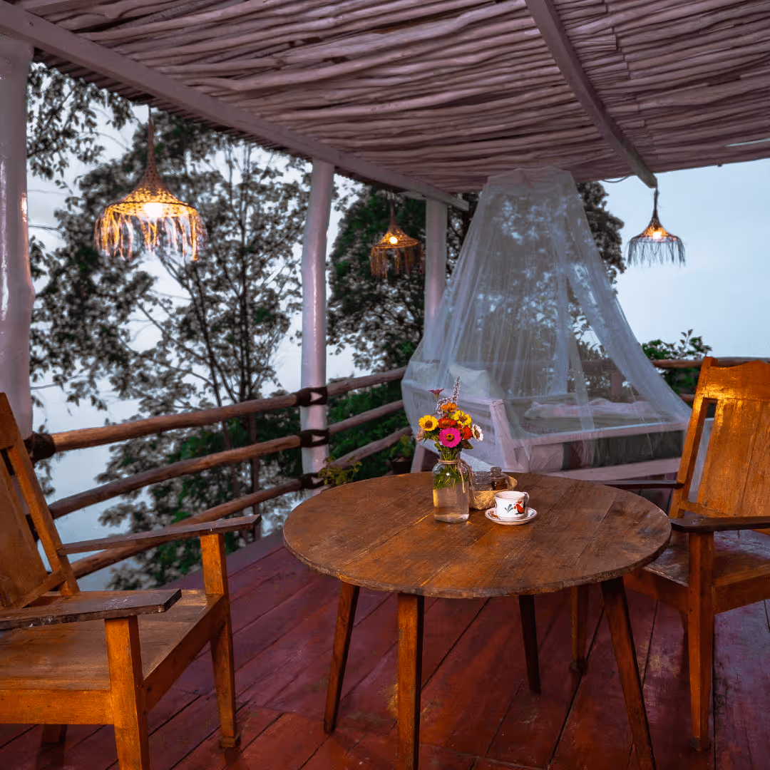 Wooden table with flowers and a cup on a covered balcony with wooden chairs, a bed with mosquito net, and hanging pendant lights amid trees.