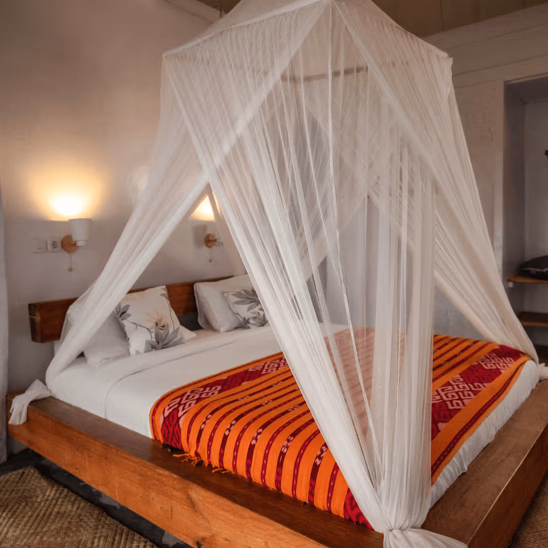Bed with wooden frame, decorative pillows, orange-red patterned blanket, and white mosquito netting canopy in a softly lit room.