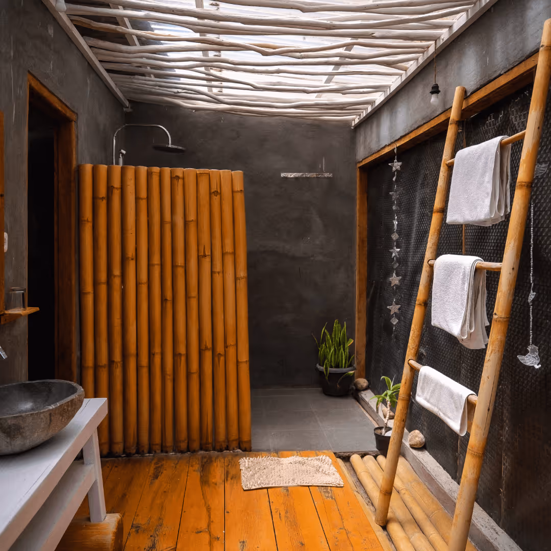 Rustic bathroom with bamboo shower divider, bamboo towel rack with white towels, stone sink, and plants on wooden floor under a pergola ceiling.