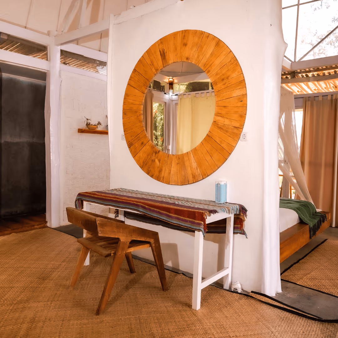 Interior room with a wooden chair, a white table covered with a multicolored cloth, and a large round wooden-framed mirror on a white wall reflecting a doorway and yellow curtain.