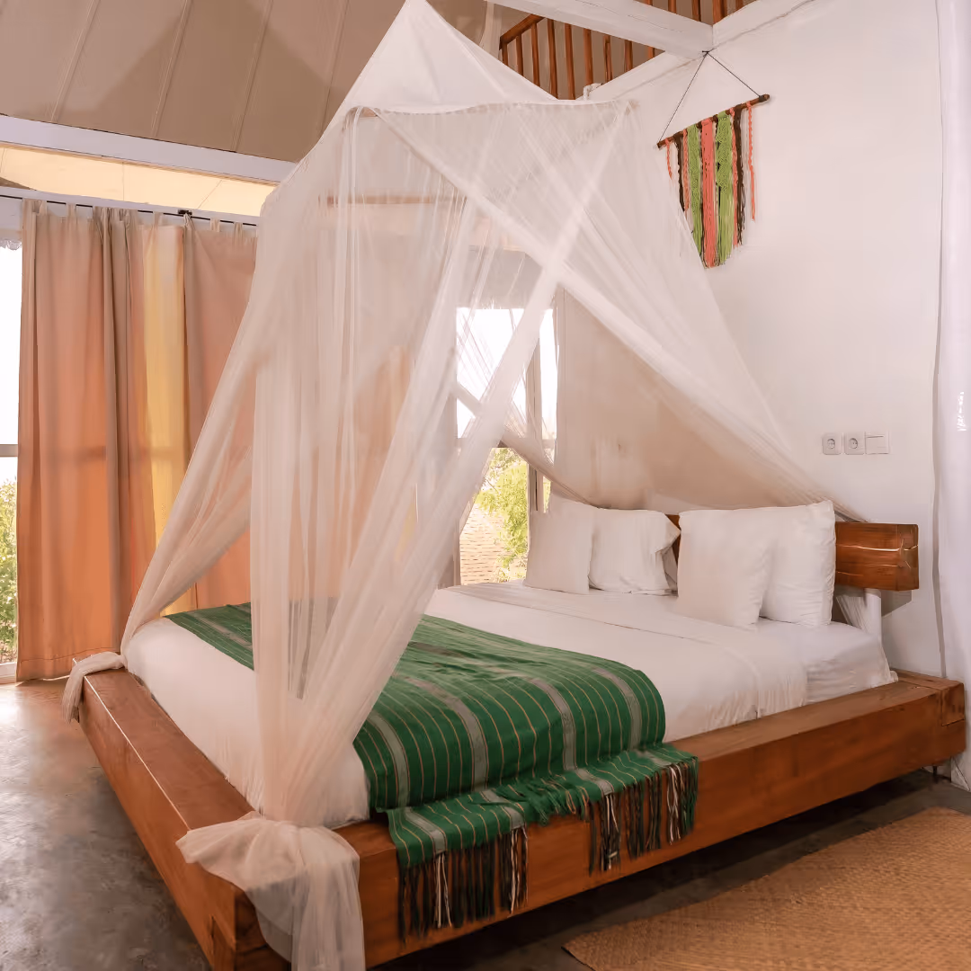 Wooden bed with white bedding, green striped throw, and a white mosquito net canopy in a bright room with beige curtains and a wall hanging.