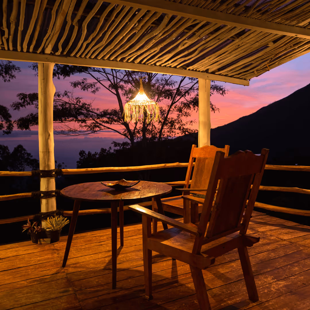 Wooden chairs and round table on a porch with a hanging light, overlooking trees and a purple-pink sunset sky.