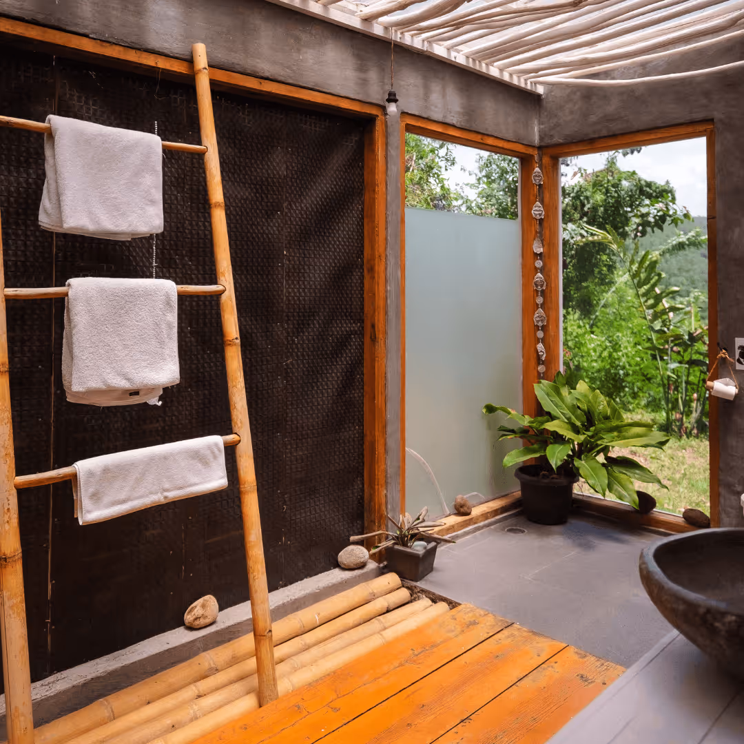 Modern bathroom corner with bamboo towel rack holding white towels, potted plants, and large windows showing green outdoor foliage.