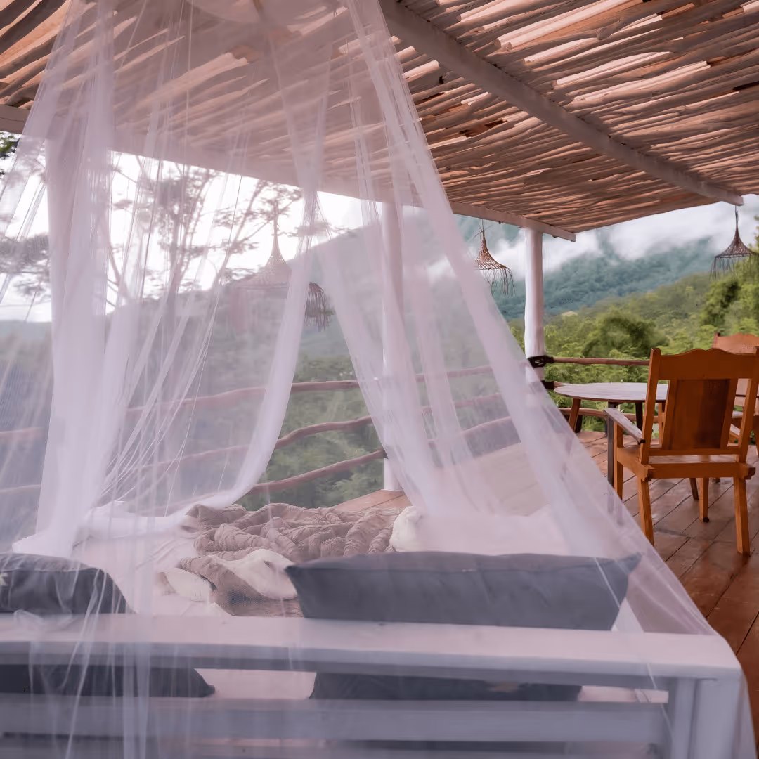 Outdoor wooden deck with a canopy bed draped in sheer white curtains, overlooking a misty mountain landscape with green trees.