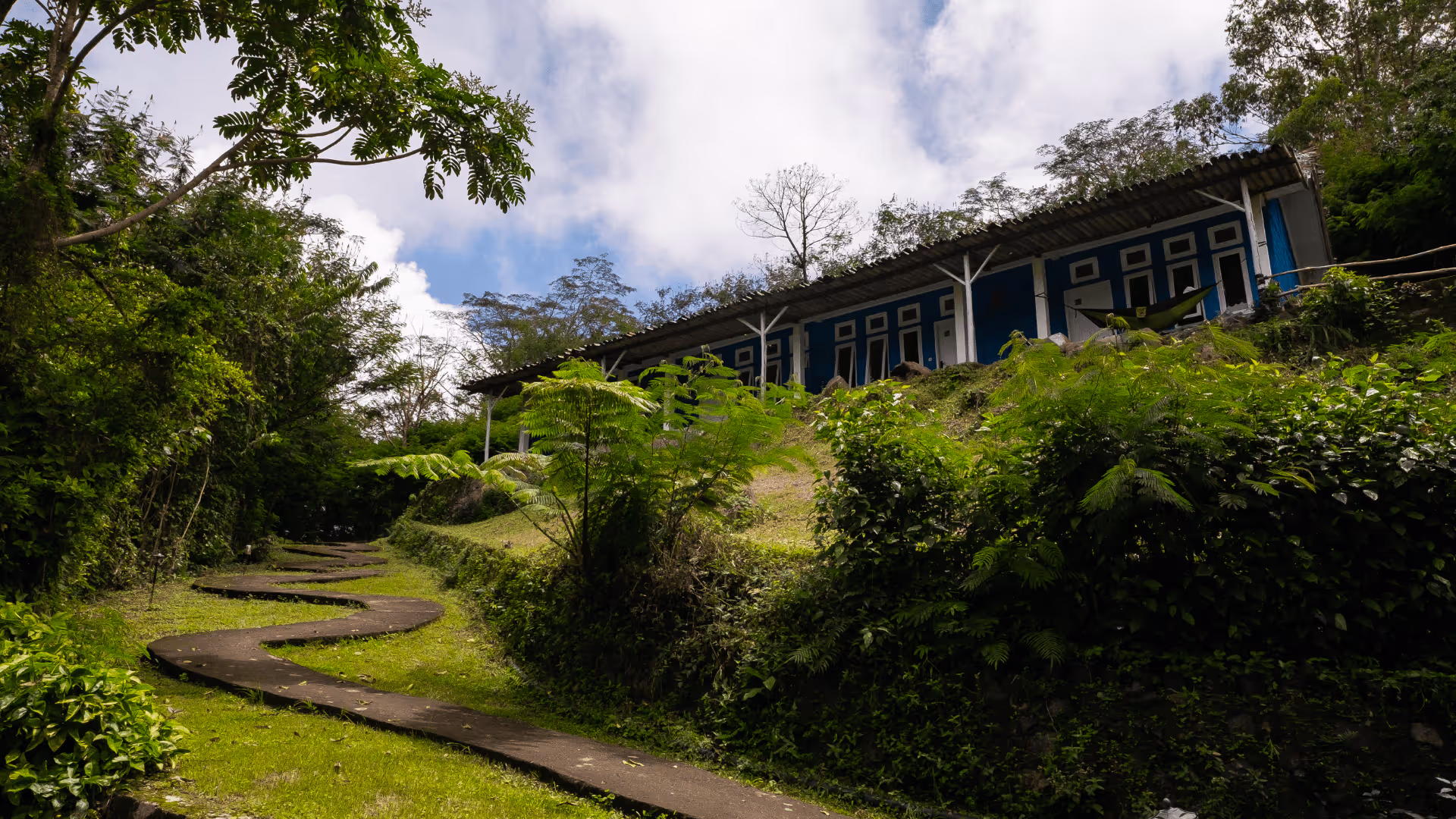 Winding paved path through lush greenery leading to a blue building with multiple doors on a hillside under a cloudy sky.