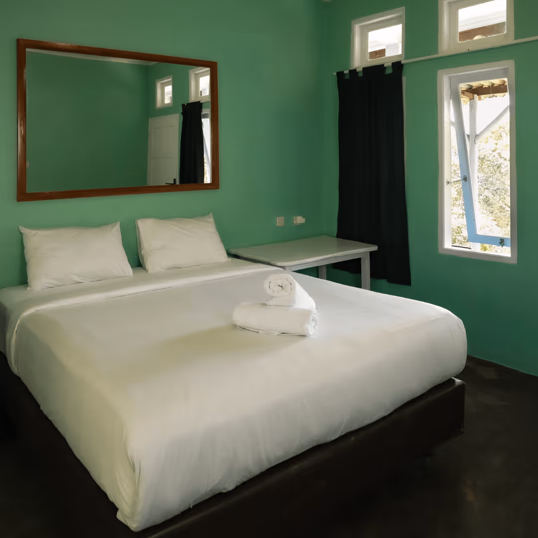 Neat bedroom with a large bed having white sheets and pillows, a pair of rolled white towels on the bed, green walls, a wooden-framed mirror, an open window, and a small table covered by a black curtain.