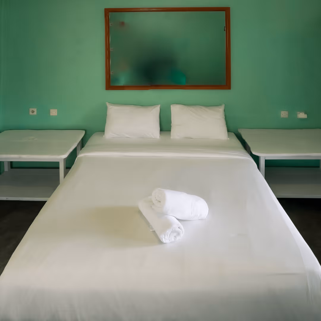 Simple bed with white linens, two pillows, and two rolled white towels, flanked by two small white bedside tables against a green wall with a wooden-framed mirror.