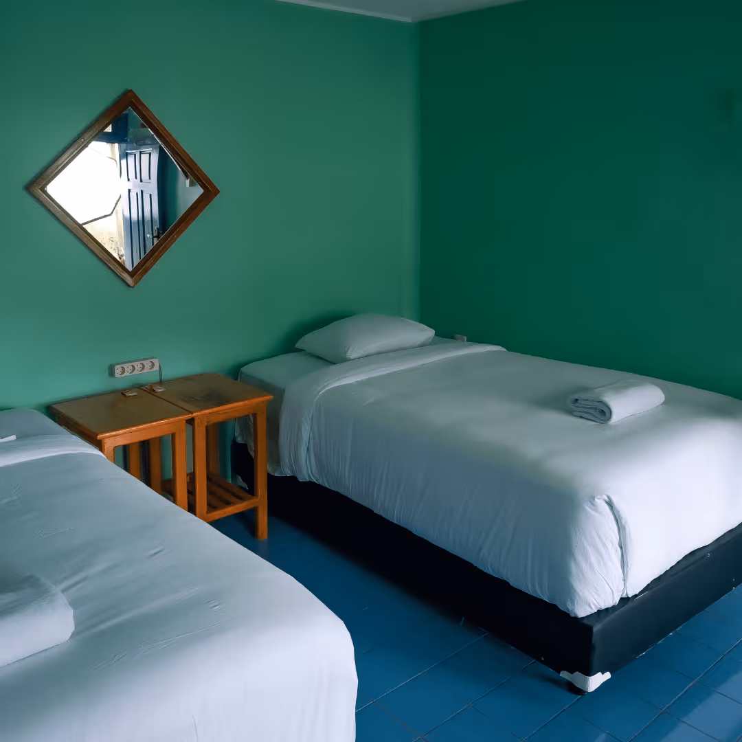 Room with two single beds featuring white linens, each with a rolled towel on top, a wooden side table between them, and a diamond-shaped mirror on green walls.