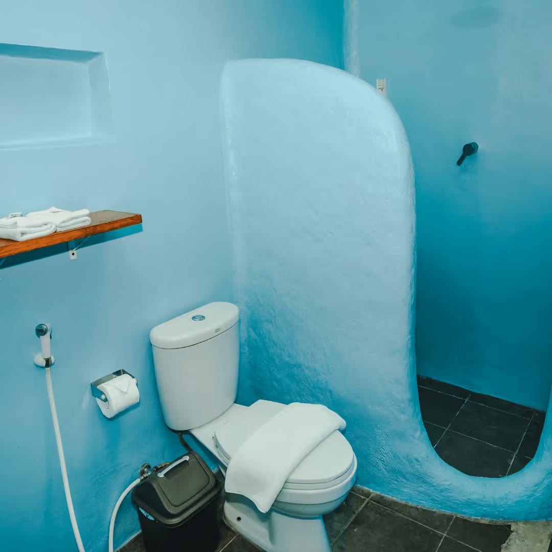 Small blue bathroom with a toilet, white towel on the lid, a wooden shelf with towels, a black trash bin, and a curved partial wall separating the shower area.