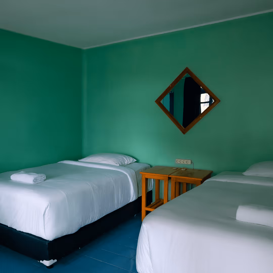 Minimalist room with two single beds with white linens, a wooden nightstand, and a diamond-shaped mirror on a green wall.