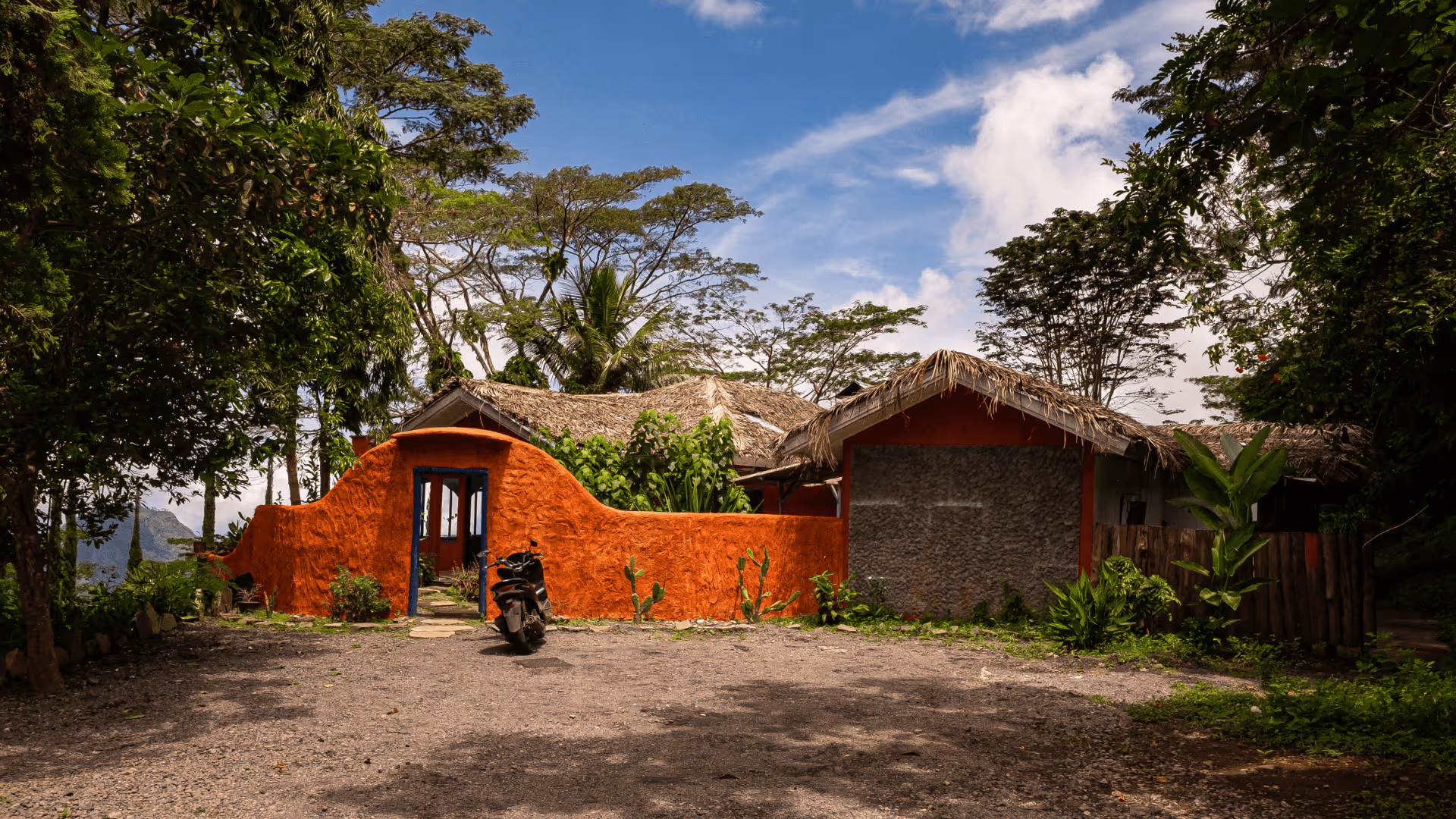 Rustic house with orange stucco walls and thatched roofs surrounded by greenery and trees under a partly cloudy blue sky.