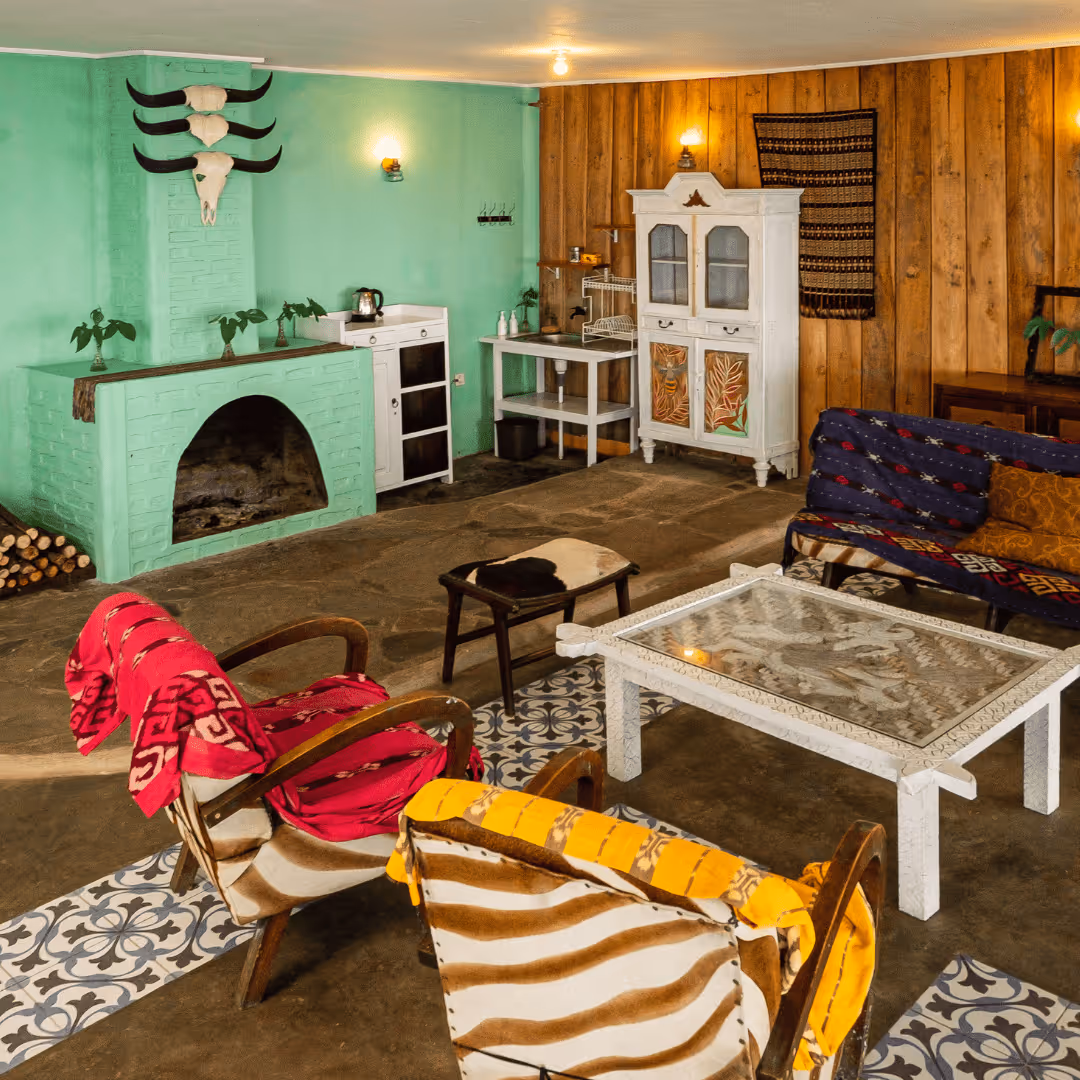 Cozy living room with green brick fireplace, patterned armchairs draped with colorful blankets, a white decorative table, and rustic wooden walls.