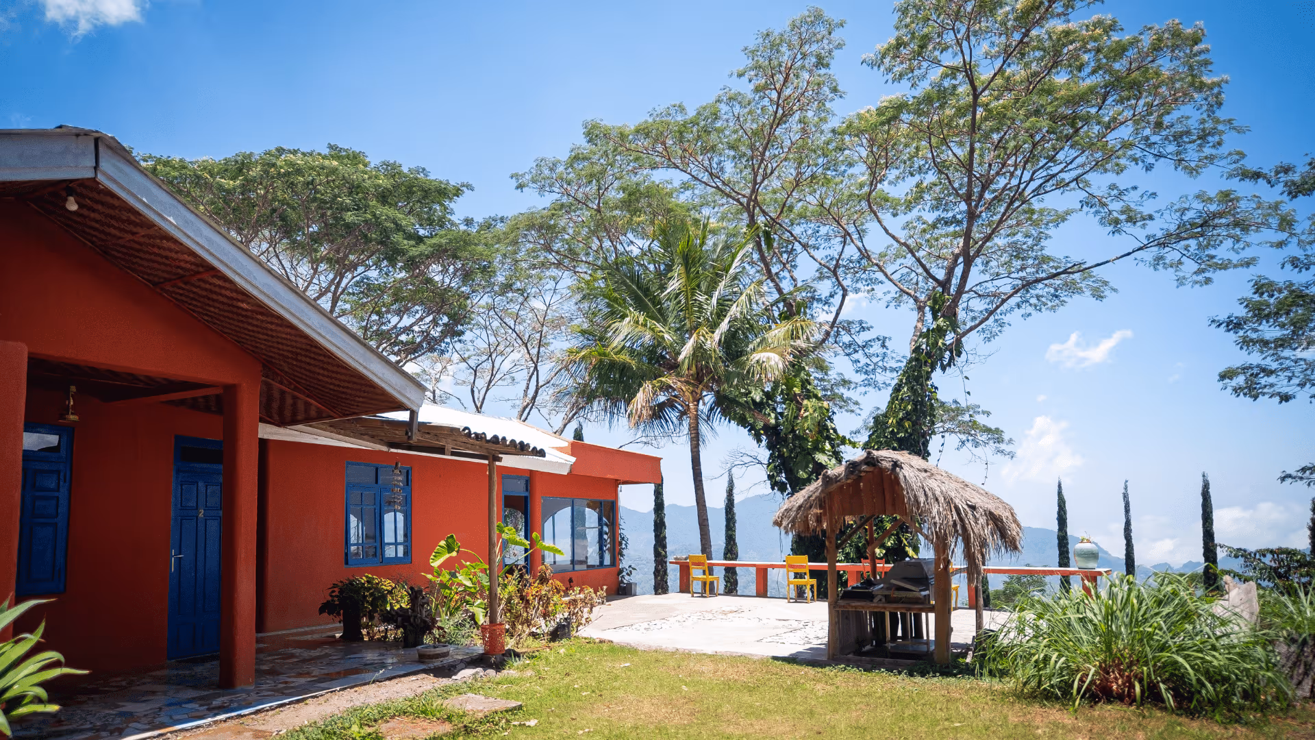 Red building with blue doors and windows next to a palm tree and thatched gazebo on a sunny day with mountains in the background.