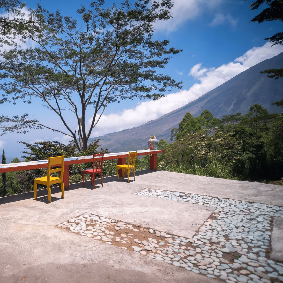 Outdoor seating area with three colorful chairs and a long bench overlooking a mountain and lush greenery under a blue sky.