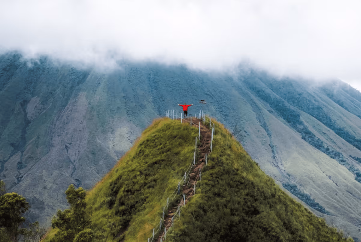 Person in red jacket with arms outstretched standing on top of a green hill with a steep, fenced path and fog-covered mountains in the background.