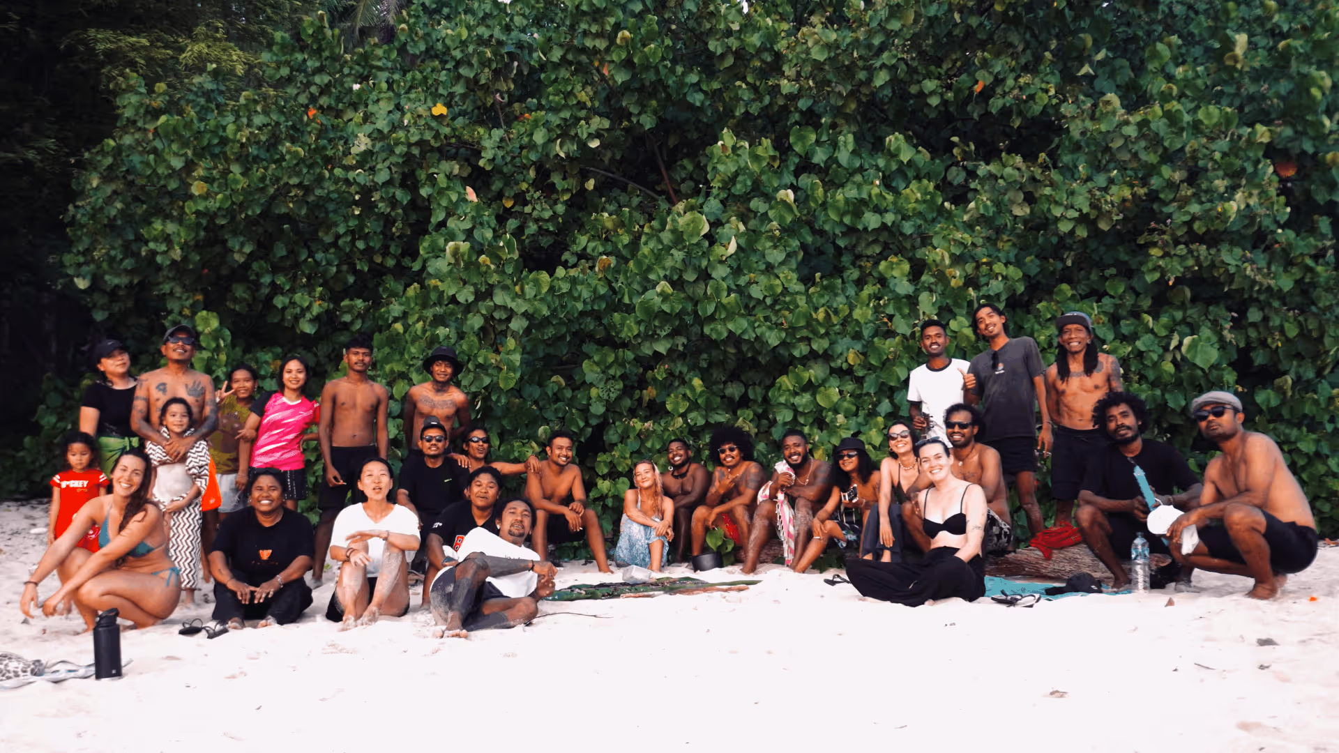 Large group of diverse people posing and sitting on sandy beach with dense green foliage background.