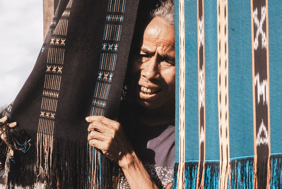 Older person peeking from between two hanging traditional woven fabrics with intricate patterns.