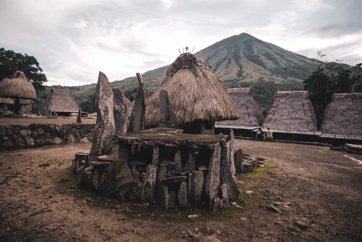 Traditional thatched huts and stone structures in a rural village with a mountain in the background.