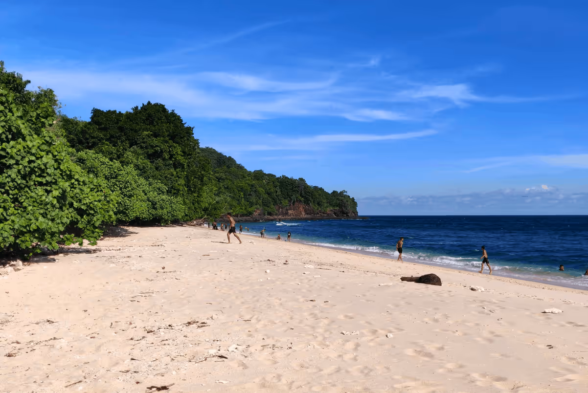 Sandy beach with people near the shoreline, lush green trees on the left, and a blue sky with wispy clouds above the ocean.