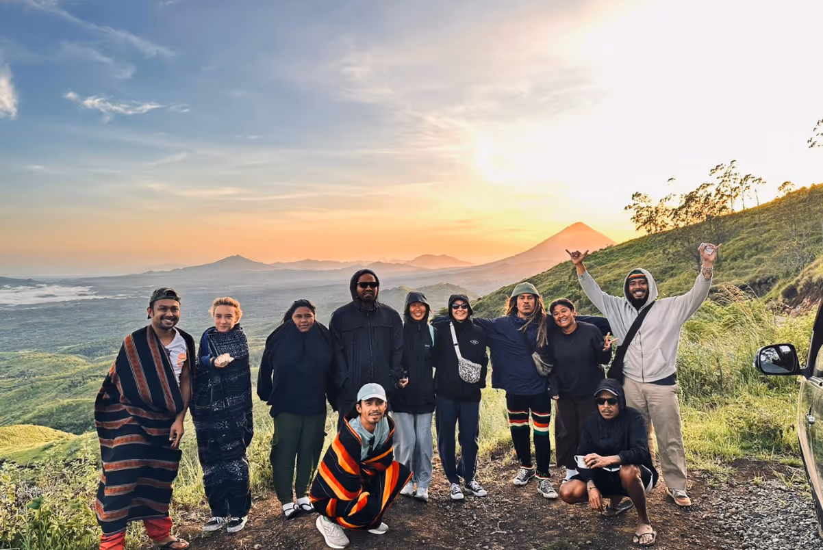 Group of ten people posing outdoors on a hillside with a scenic sunset and mountains in the background.