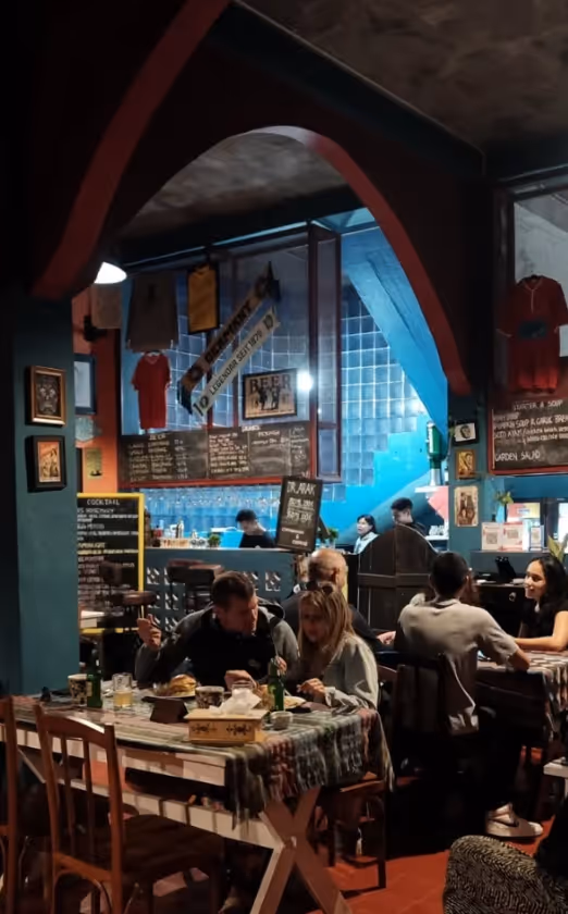 Cozy indoor bar with people sitting at tables enjoying food and drinks, colorful tiled walls and a menu board behind the bar.