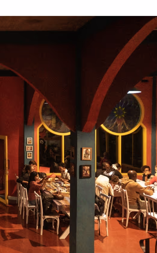 Group of people sitting around two tables engaged in conversation inside a warmly lit room with arched ceilings and large round windows.