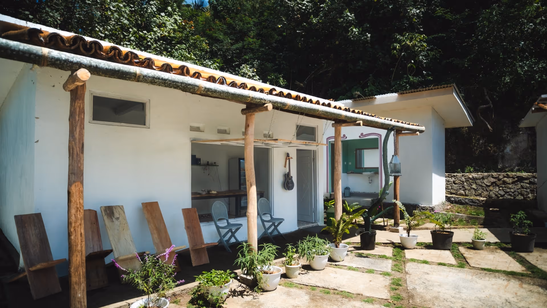 White building with a tiled roof, wooden chairs and potted plants under a covered porch surrounded by greenery.