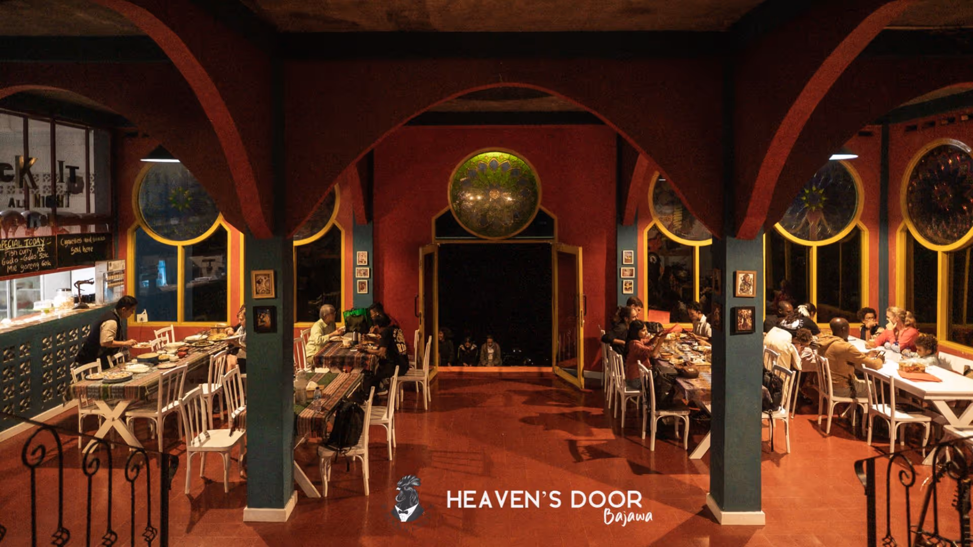Interior of Heaven's Door Bajawa restaurant with diners seated at tables under arched ceilings and round stained glass windows.
