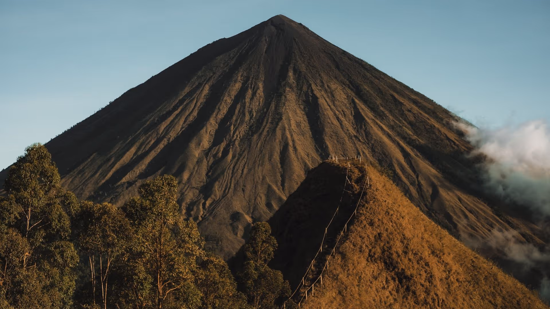 Tall conical volcanic mountain with a smaller hill in front, trees at the base, and partly cloudy sky.