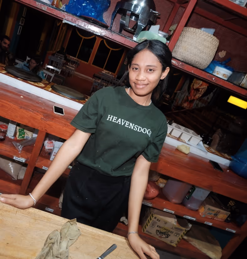 Young woman wearing a dark green HEAVENSDOC t-shirt standing behind a wooden counter in a shop or kitchen setting.