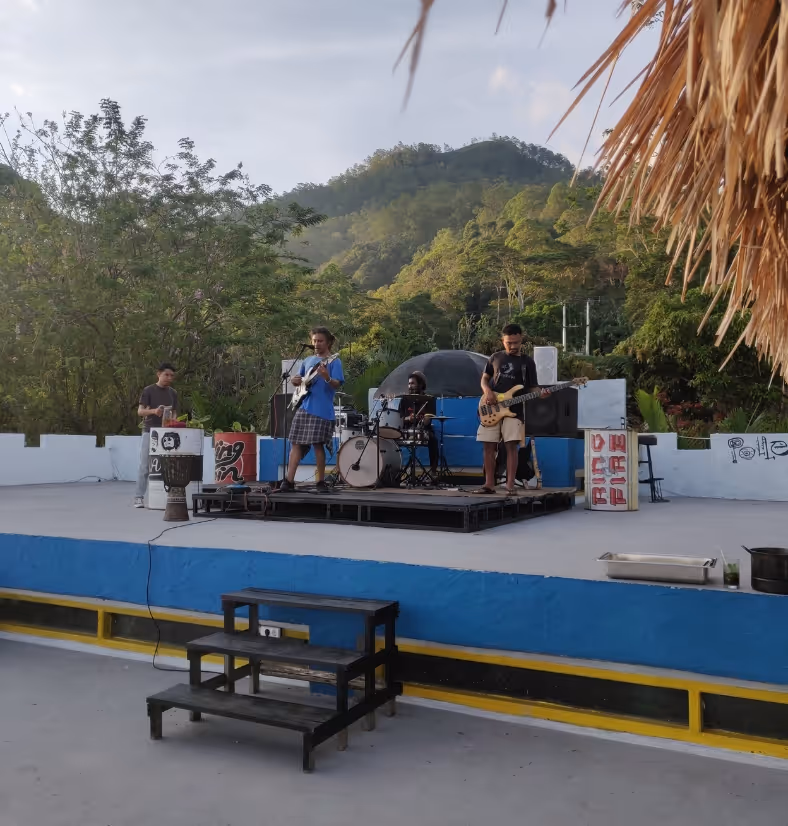 Outdoor band performing on a small stage with guitars and drums, surrounded by trees and hills in the background.