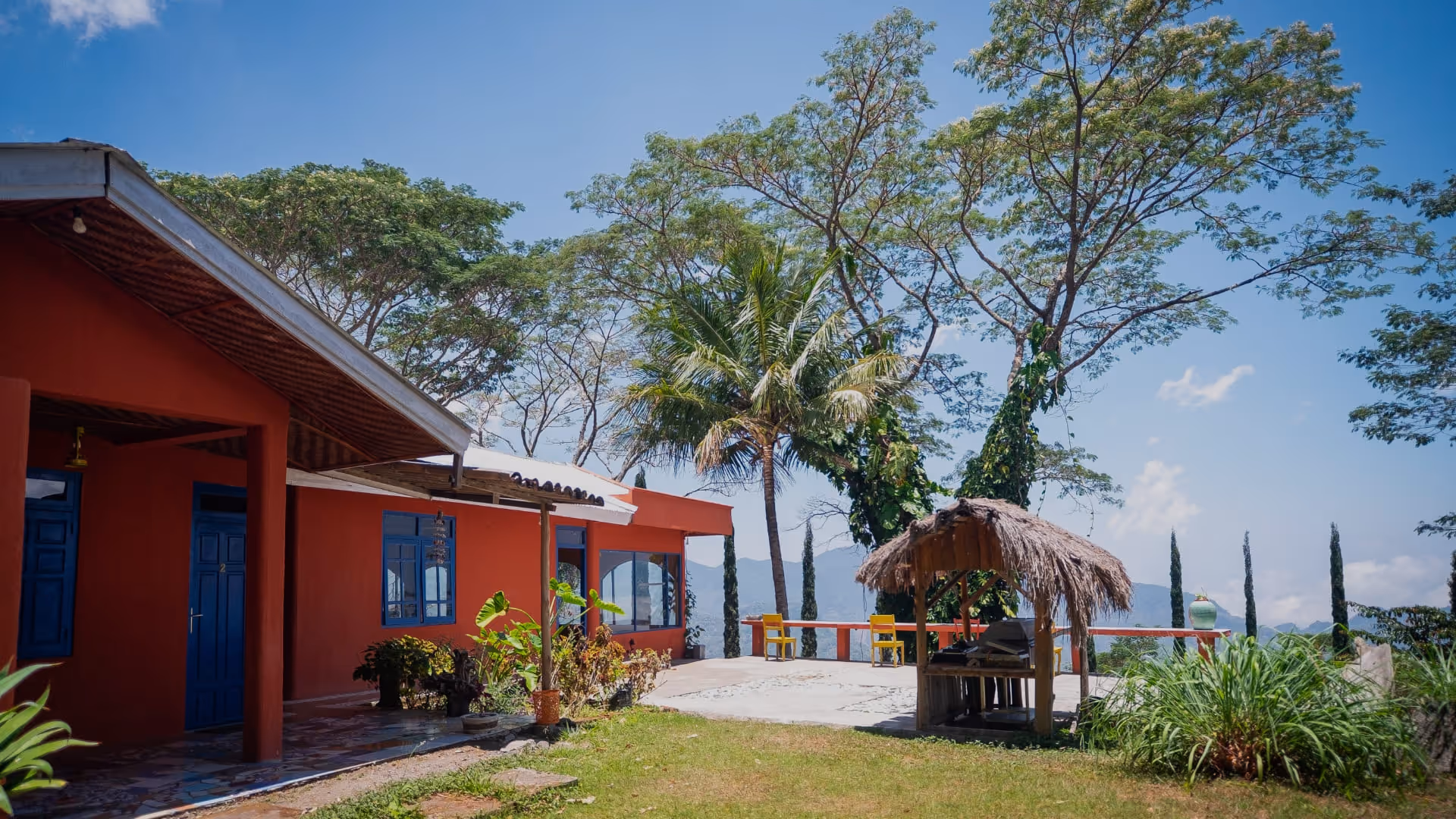 Red building with blue doors and windows next to a patio, tropical palm trees, and a palapa-style shaded seating area under a clear blue sky.