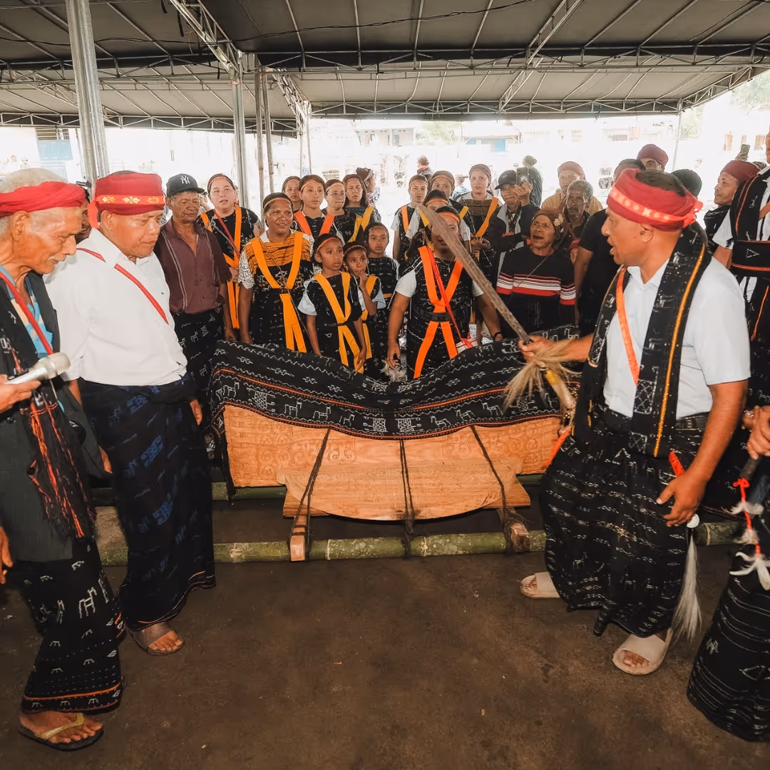 Group of people dressed in traditional attire gathered around a carved wooden coffin under a canopy, participating in a cultural ceremony.