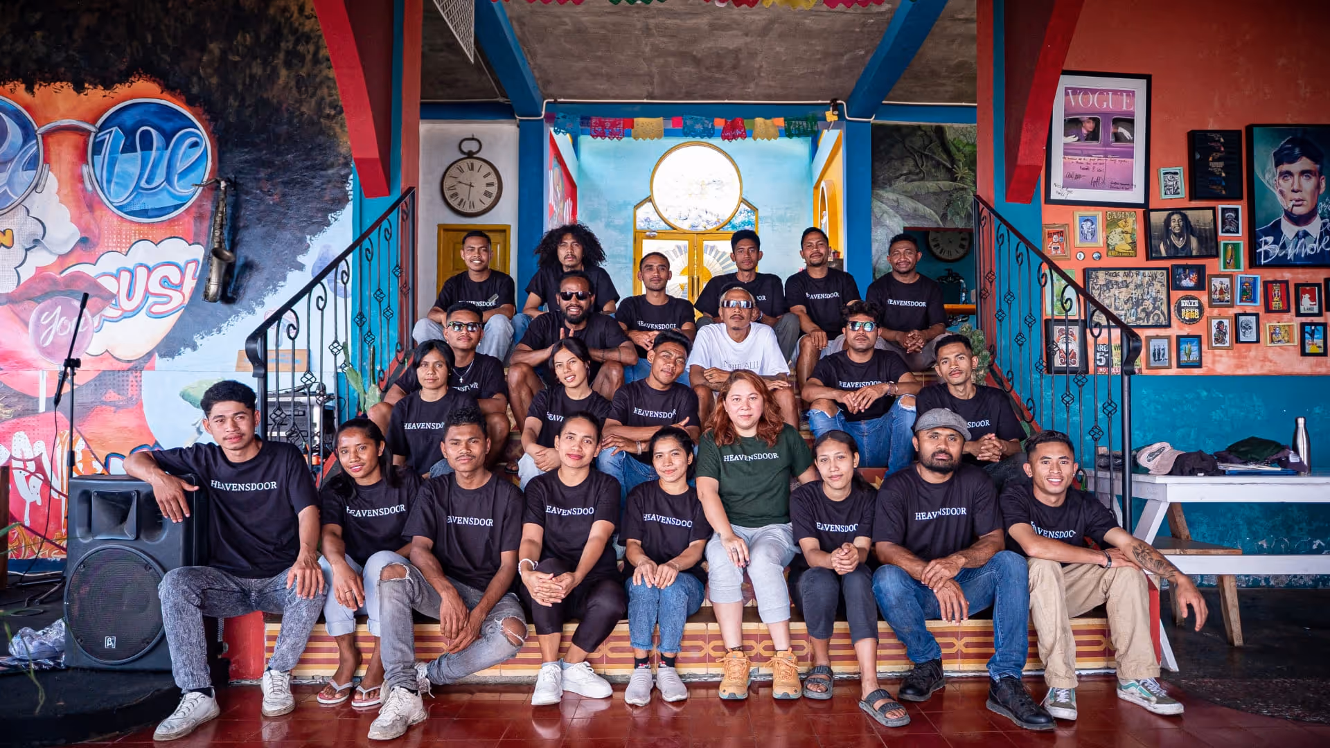 Group of people wearing black and green 'HEAVENSDOOR' t-shirts sitting on steps inside a colorful room with wall art and framed pictures.