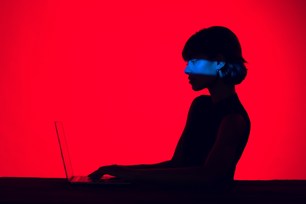 A person sits in profile at a laptop, silhouetted against a solid red background, with a strip of blue light across their face.