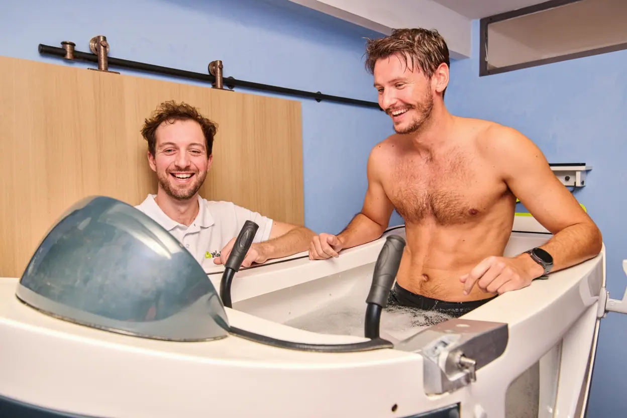 Un homme torse nu souriant est dans une machine d'aquabiking tandis qu'un autre homme, habillé d'un polo blanc, le regarde et sourit.