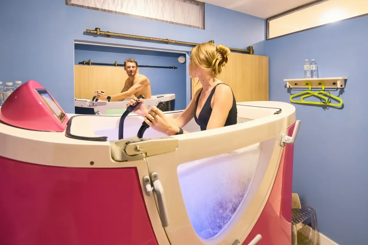 Une femme pratique du aquabiking dans une cabine équipée d'un vélo aquatique rose, regardant un homme en reflet dans un miroir dans une salle avec des murs bleus.