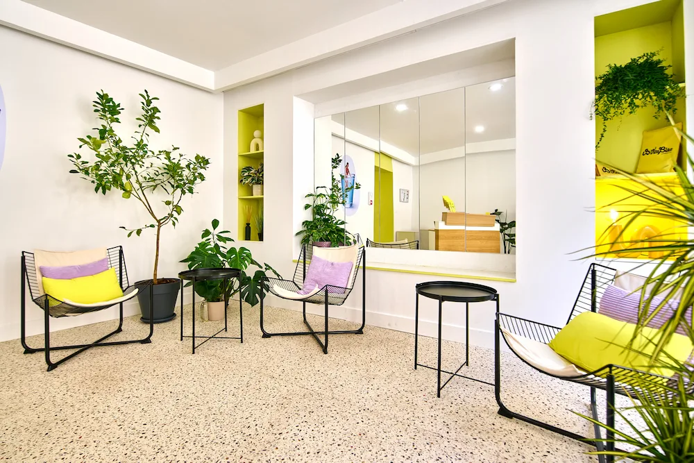 Coin salon moderne avec trois fauteuils noirs en métal, coussins jaunes et violets, deux petites tables rondes noires, plantes vertes et grand miroir mural.