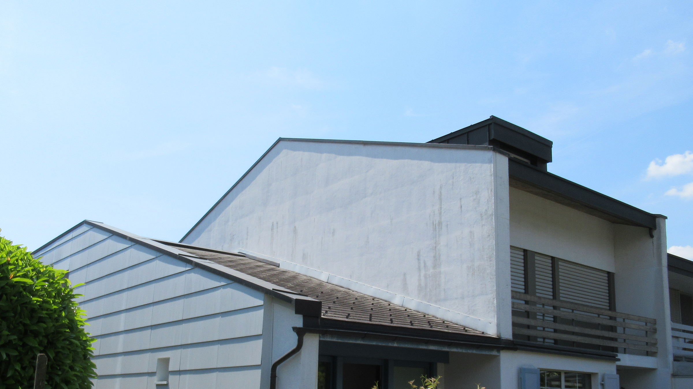 Weisses Wohnhaus mit Balkon, geschlossenen Fensterläden und dunkelgrauem Dach unter blauem Himmel.