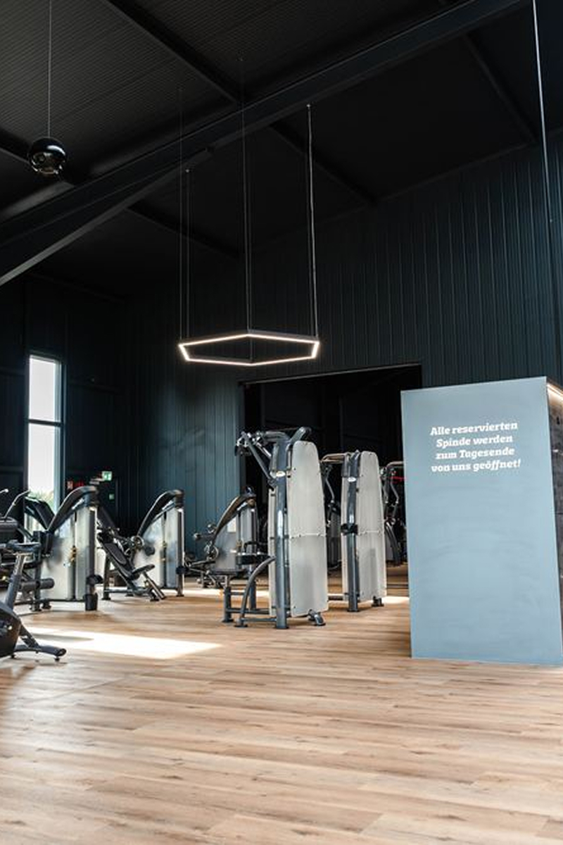 Modern gym interior with exercise machines, wooden floor, dark walls, and a hanging hexagonal light.
