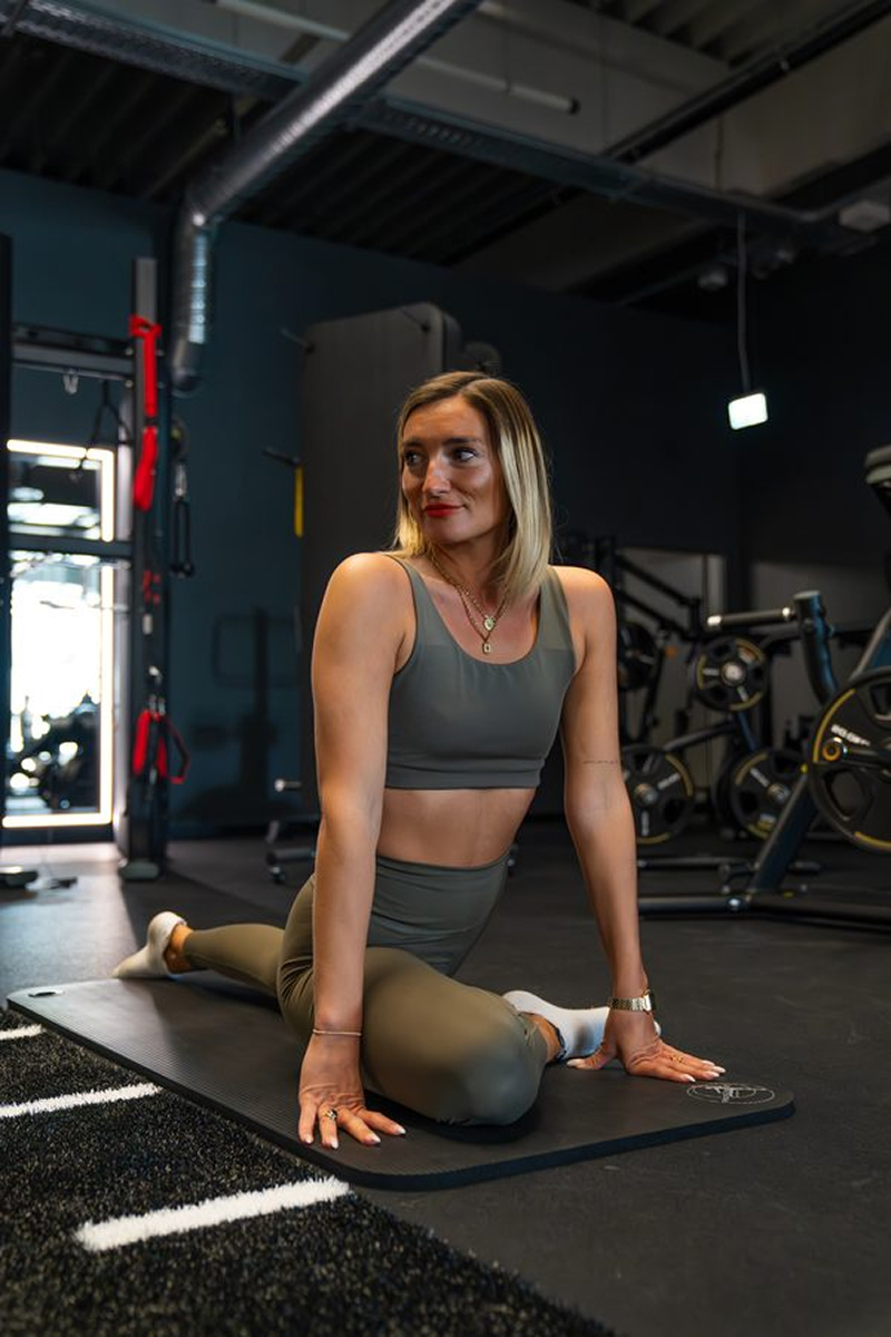 Woman in workout clothes doing a seated yoga stretch on a black mat in a gym.