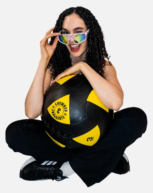 Smiling woman with curly hair sitting cross-legged holding a black and yellow ball wearing sunglasses.