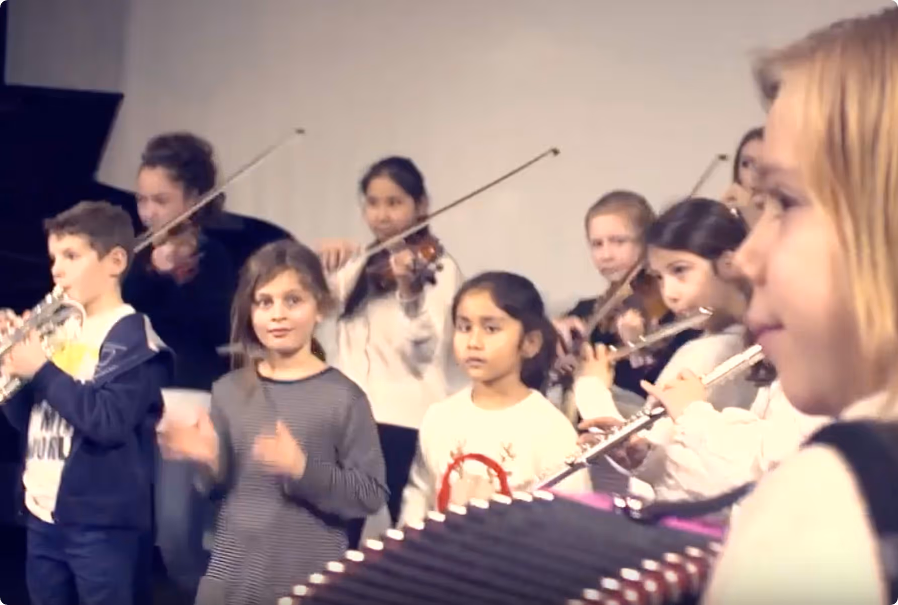 Groupe d'enfants jouant de différents instruments de musique, notamment violons, flûte et accordéon, dans une salle.