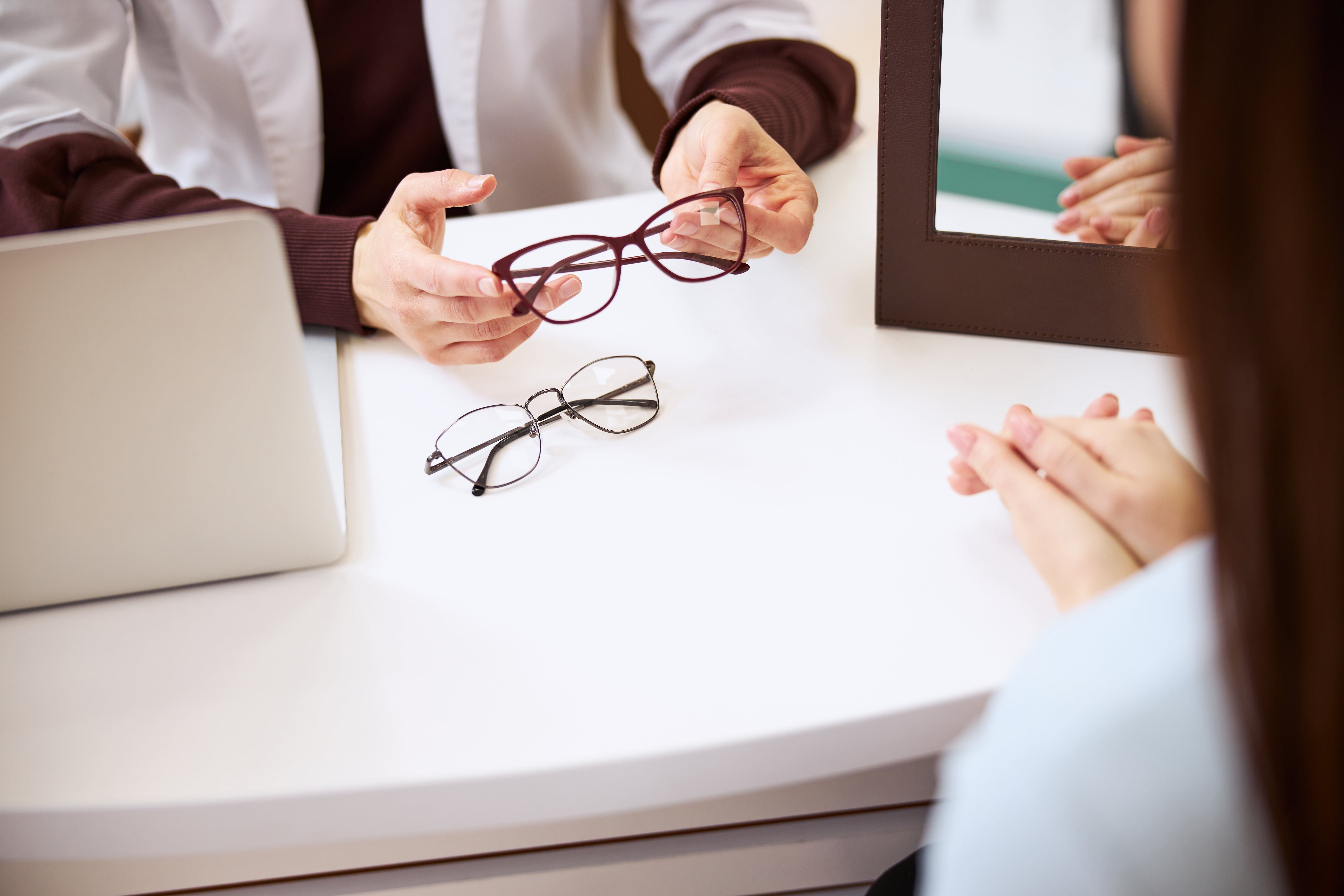 Person holding eyeglasses on white desk near laptop and hands
