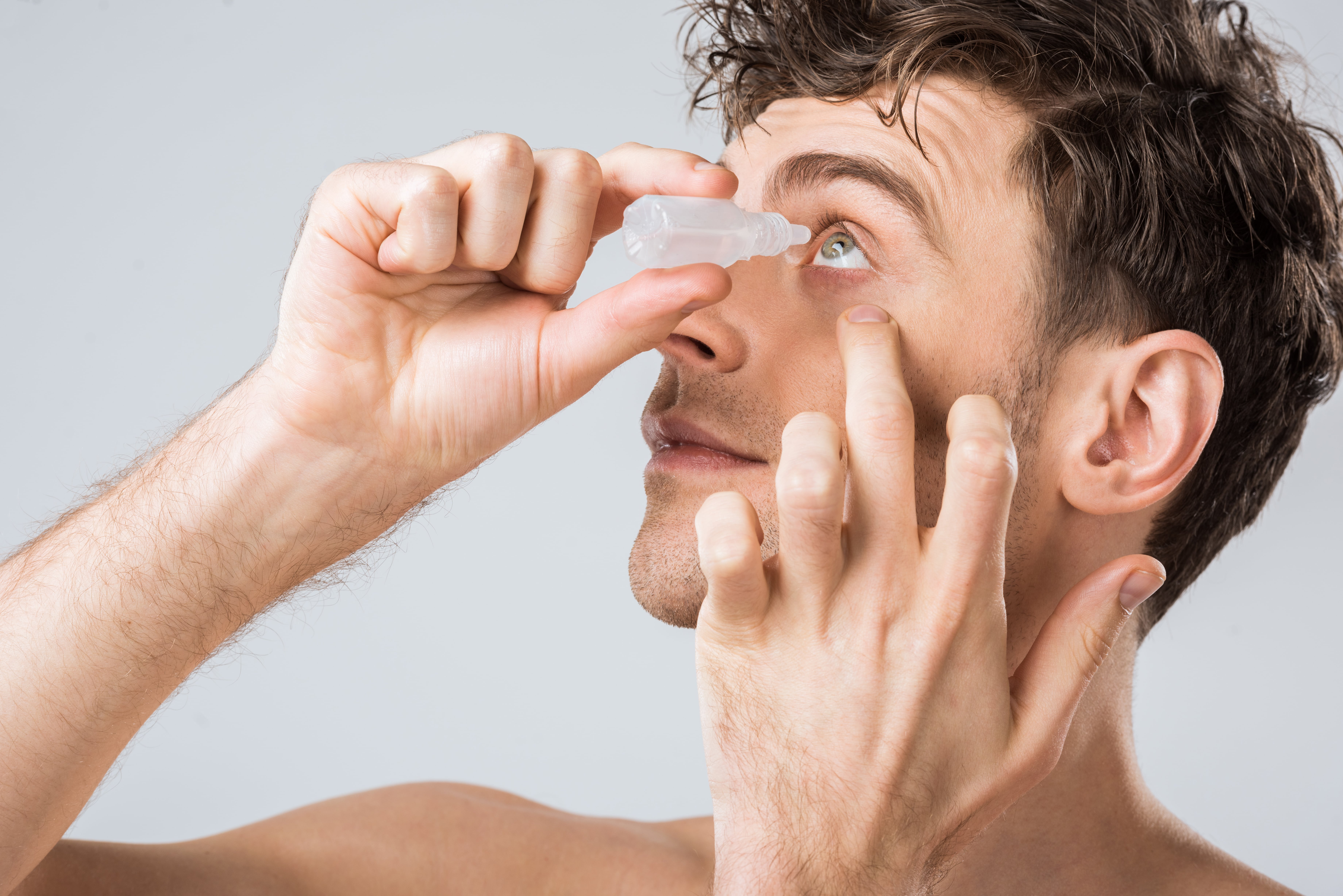 Person applying eye drops with focused expression against neutral background
