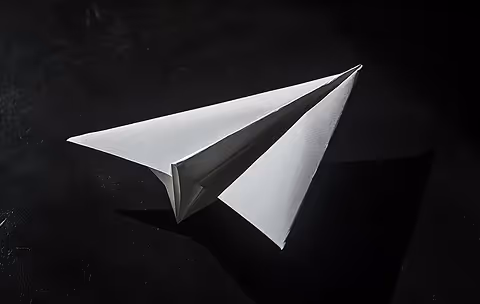 A white paper airplane resting on a dark surface, lit from above, casting a sharp shadow to the right.