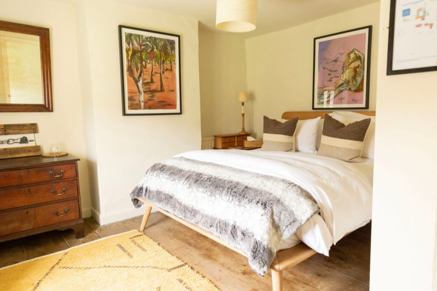 Cozy retreat like bedroom with a wooden bed, white bedding, striped grey throw, two pillows, wooden furniture, framed artwork, and a yellow rug on wooden floor.